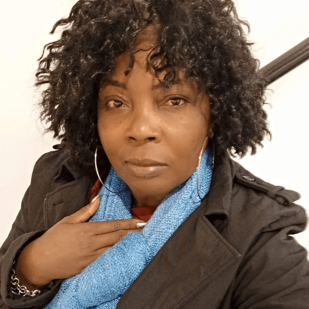 Portrait d'une femme avec des cheveux crépus, portant une écharpe bleue et une veste noire, affichant un visage sérieux et une main sur la poitrine.